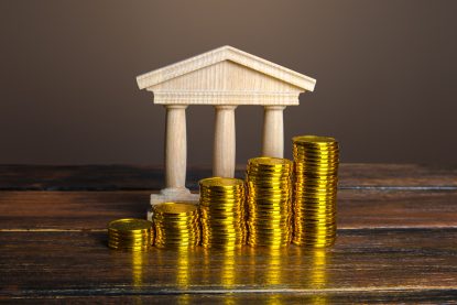 Uma pilha de moedas douradas a frente de uma miniatura de construção de uma instituição financeira